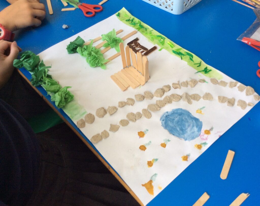 The image shows a multimedia piece on a desk in a classroom. Two children's hands and some art equipment are visible. The piece depicts a garden containing planted areas, a rock-edged path and a pond. In the centre is a wooden structure.