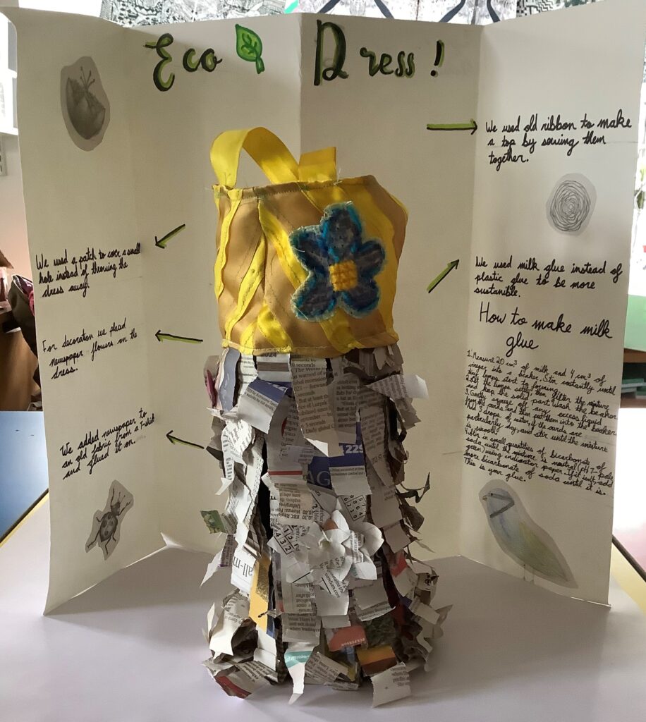 A 3D model of a dress. Its bodice is created from recycled beige and yellow ribbons, with a simple large blue flower motif. The skirt is created out of overlapping pieces of recycled newspaper fixed to fabric from an old T-shirt, and decorated with newspaper flowers. Behind the dress is an explanatory panel which includes a recipe for milk glue.